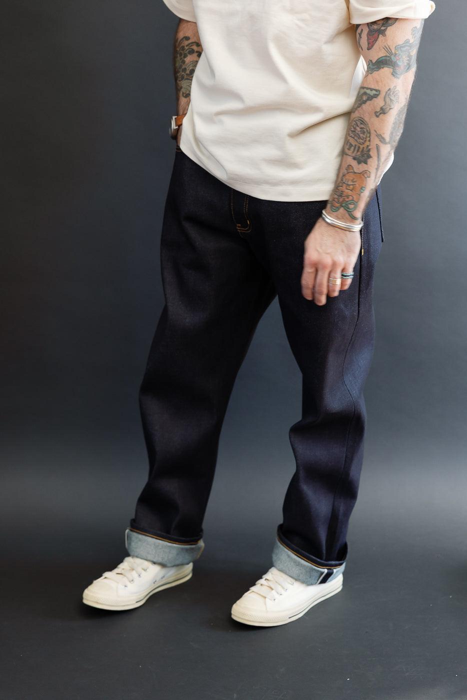 Full body front view of a male model with an athletic build wearing dark selvedge denim jeans and white sneakers.