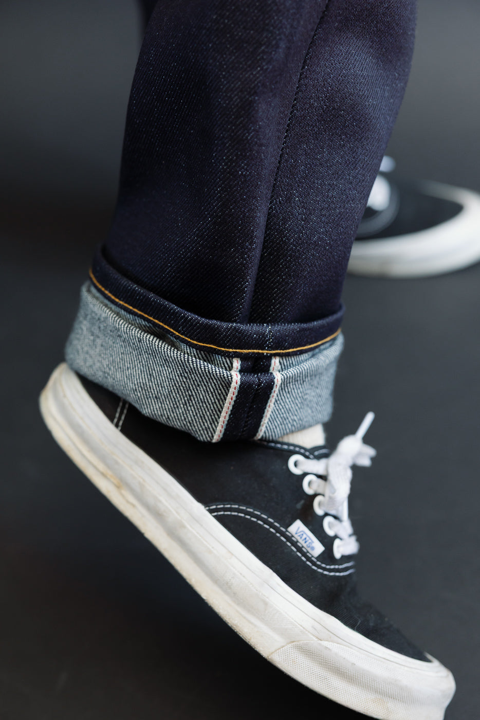 Close-up of the cuffed hem and selvedge detail on dark indigo denim jeans worn with black sneakers.