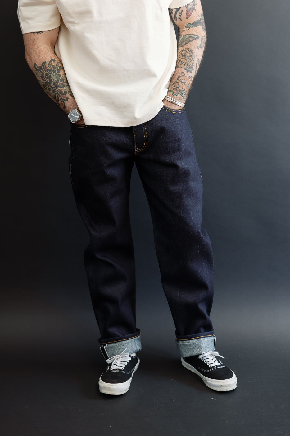 Full front view of a male model with an athletic build wearing dark indigo selvedge denim jeans and black sneakers.