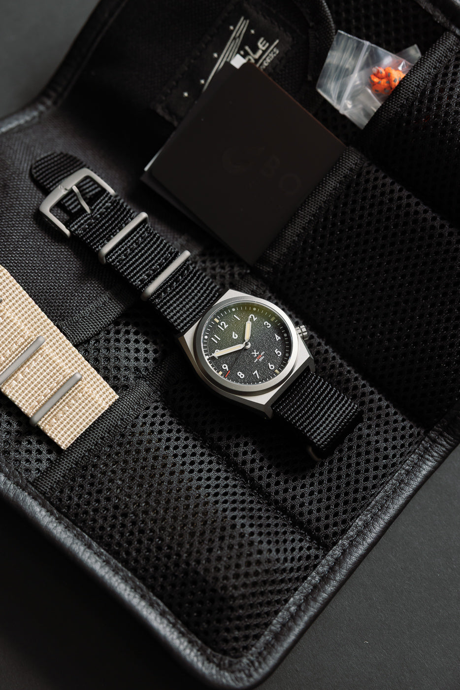 Close-up of black field watch with nylon strap in carrying case, front view of watch face and strap details.