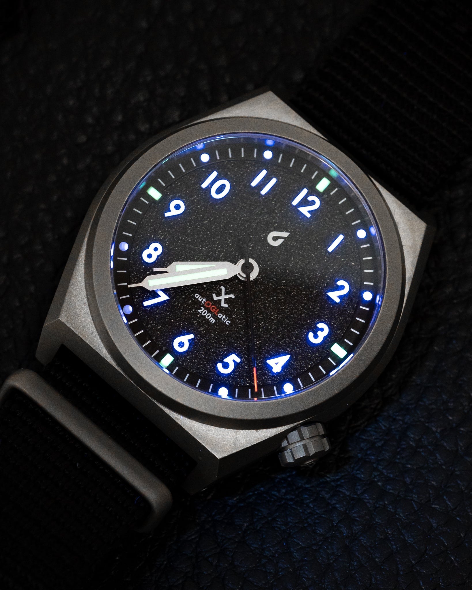 Close-up of black field watch dial with luminous blue numerals, crown, and nylon strap front view.