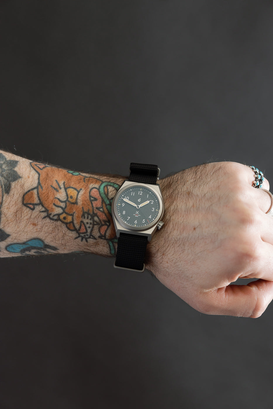 Close-up of black field watch on male model wrist, focus on watch face and band, left side view.