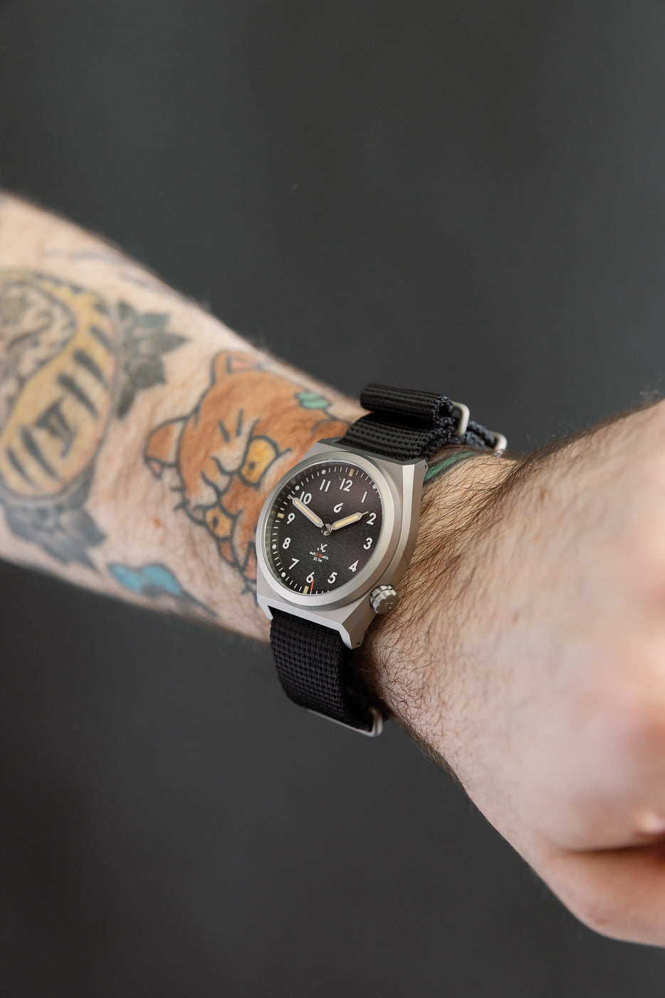 Close-up of black dial field watch with nylon strap on male model's wrist, side view.