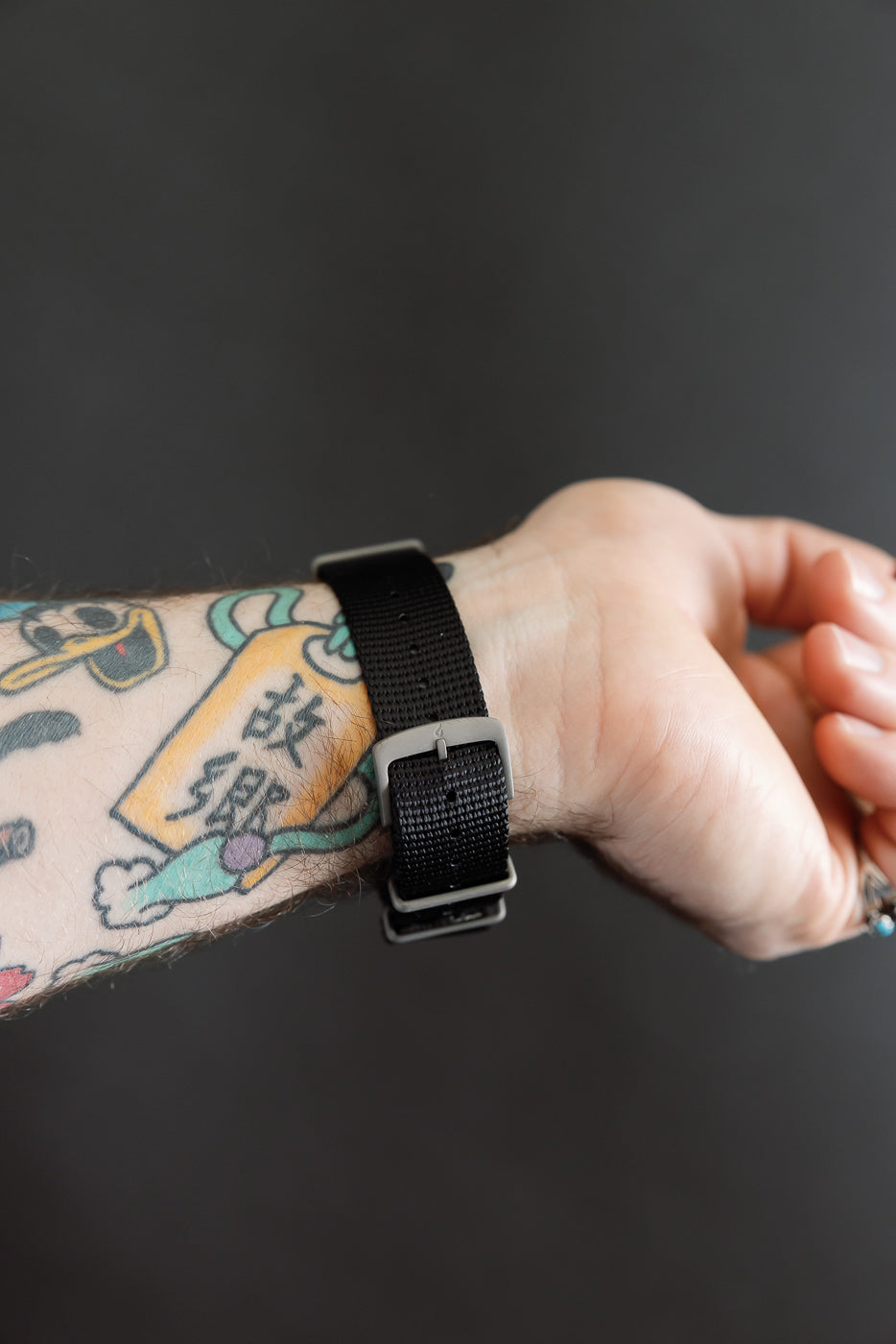 Close-up of black nylon watch strap with silver buckle on male model’s wrist, back side view