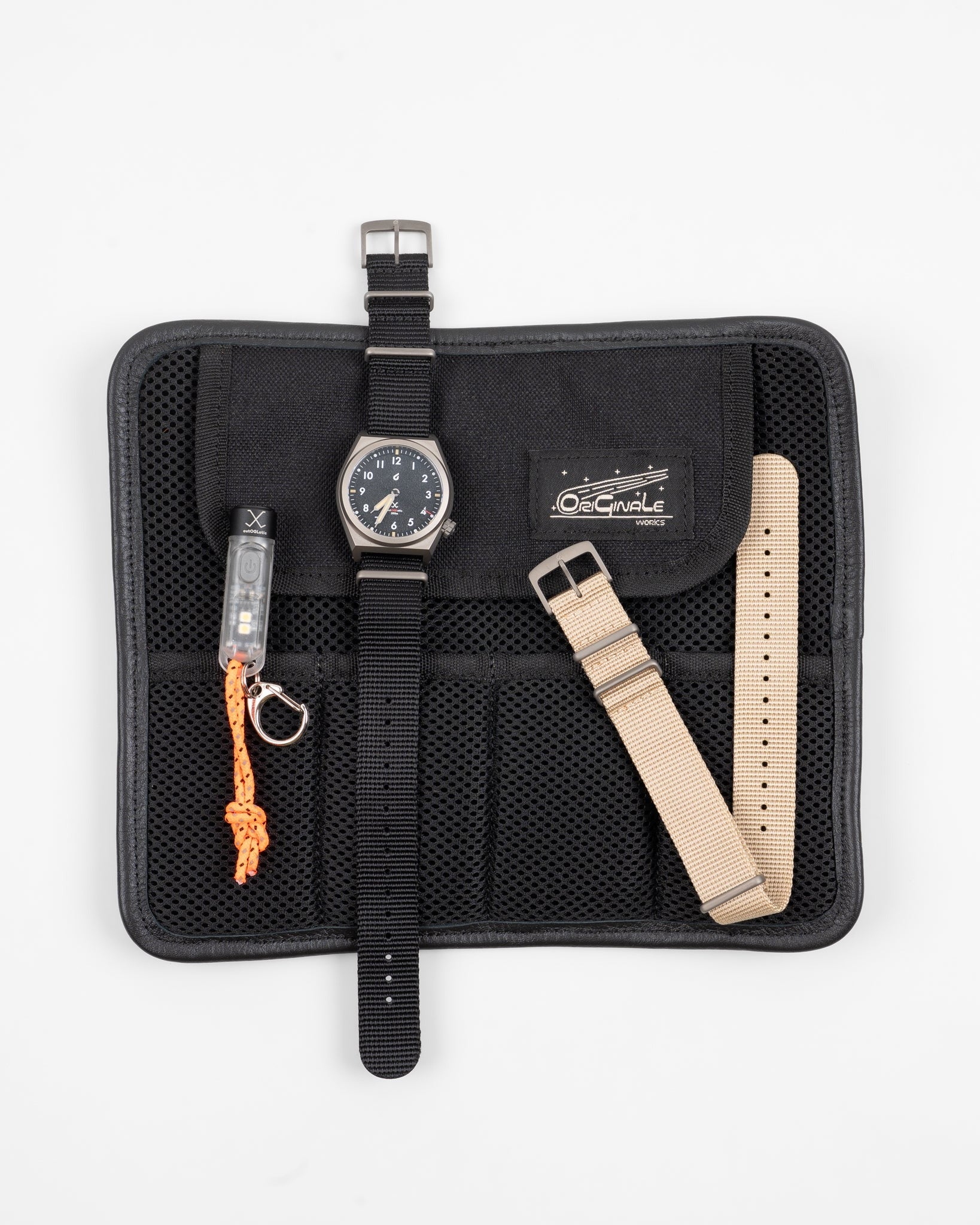 Flat lay close-up of black field wristwatch with black nylon strap, additional beige strap, and watch carrying case showing front side details and accessories.