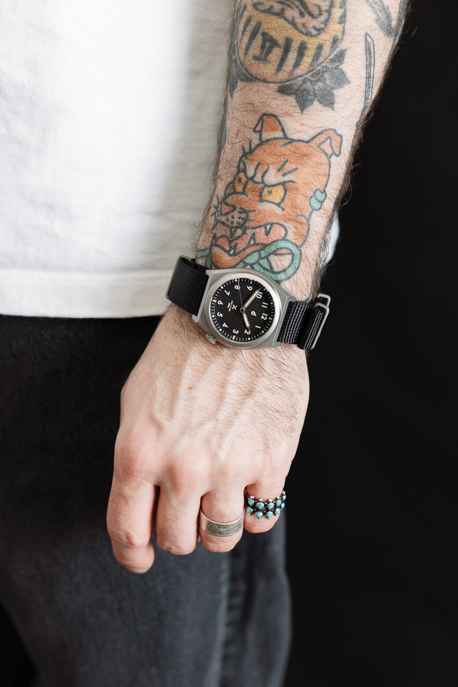 Close-up of male model's left wrist with black field watch, tattoo and rings visible, partial side view.