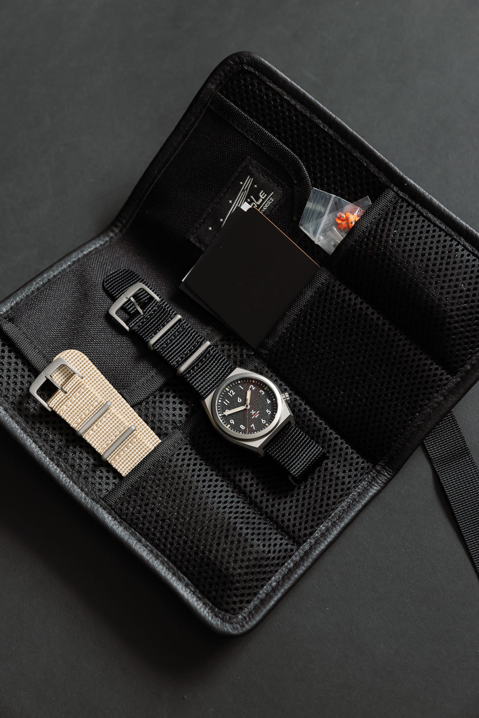 Close-up of field watch with black and tan nylon straps, laid flat in open mesh travel case, front view.