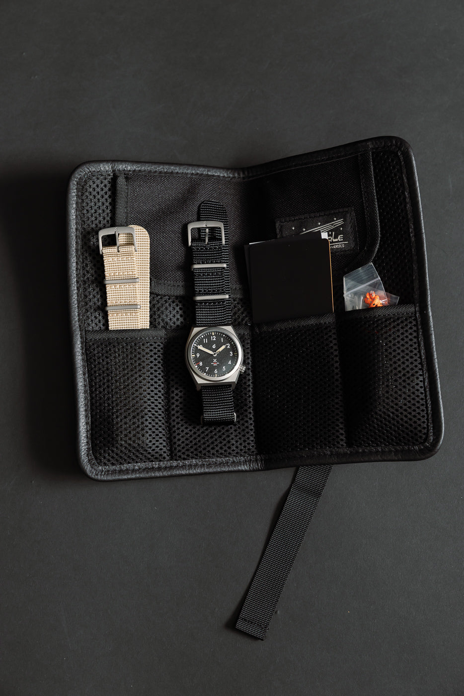 Flat lay close-up of front side: field watch with black strap, beige strap, mesh travel case.