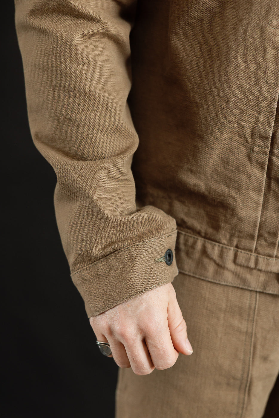 Close-up of a light brown 01507-HOX-BRK - 13oz Brown Khaki Sulfur Dyed Heavy Oxford Jacket, showcasing the buttoned cuff and textured fabric, complemented by a silver ring on the hand.