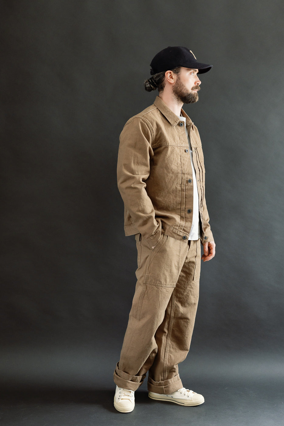 Man in profile wearing 01507-HOX-BRK - 13oz Brown Khaki Sulfur Dyed Heavy Oxford Jacket - Type I, with double pleats, copper rivets, and cinch back, styled with matching trousers and sneakers.