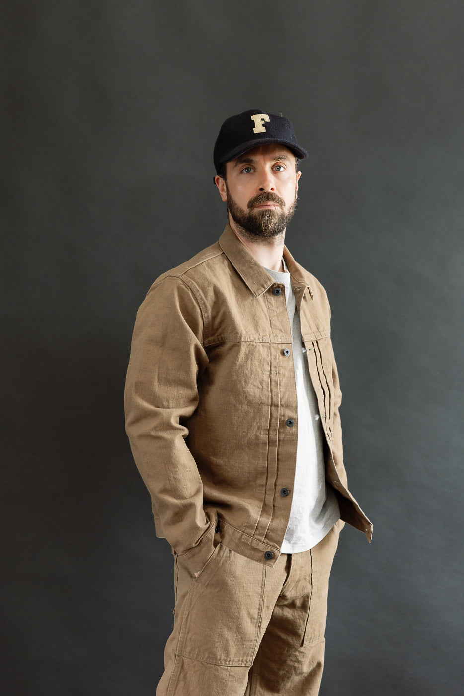 A man models the 01507-HOX-BRK - 13oz Brown Khaki Sulfur Dyed Heavy Oxford Jacket - Type I, showcasing its classic workwear silhouette, single chest pocket, and distinct pleating.