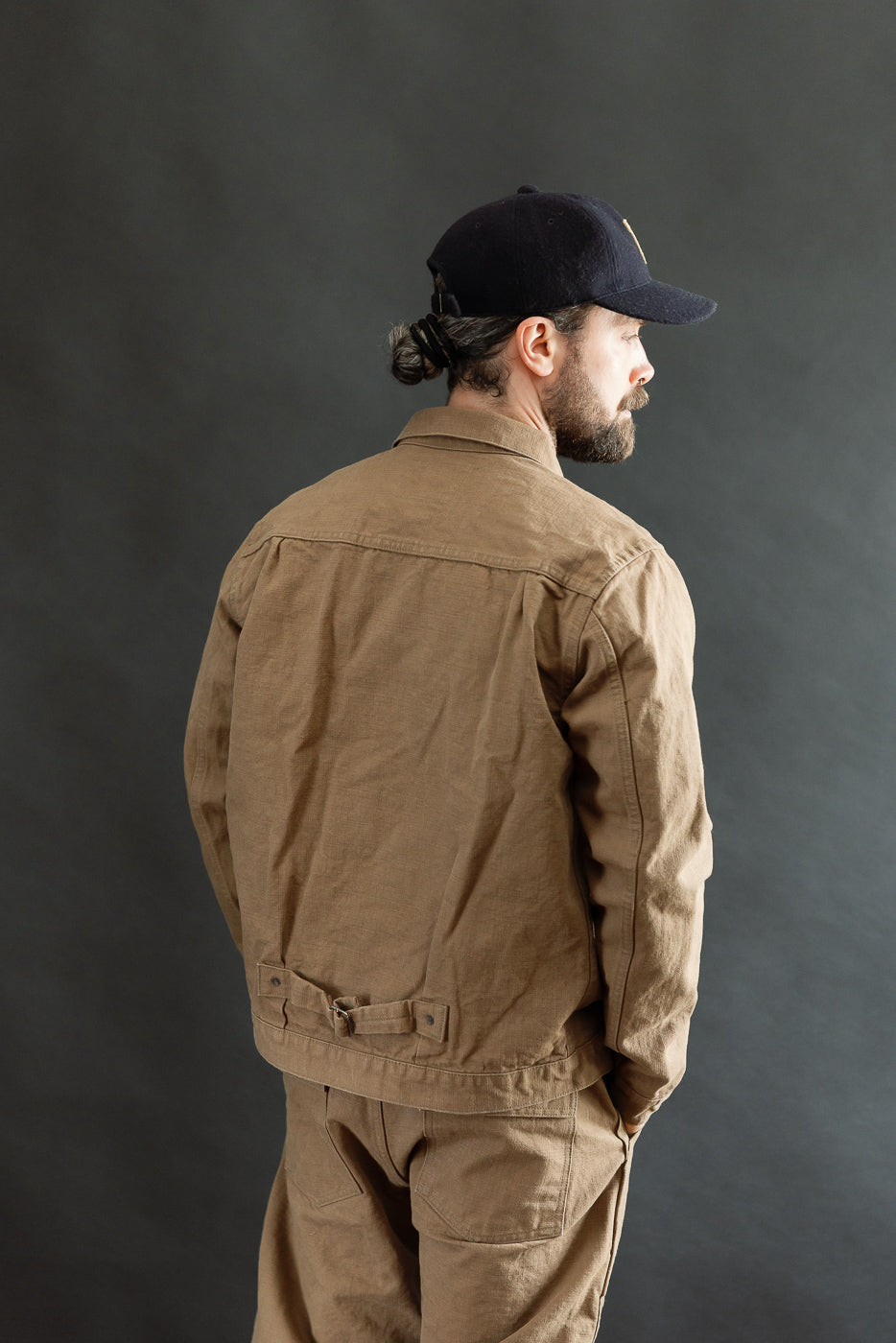 A man models the ONI Denim 01507-HOX-BRK - 13oz Brown Khaki Sulfur Dyed Heavy Oxford Jacket - Type I, showcasing its workwear style with a cinch back and single chest pocket.