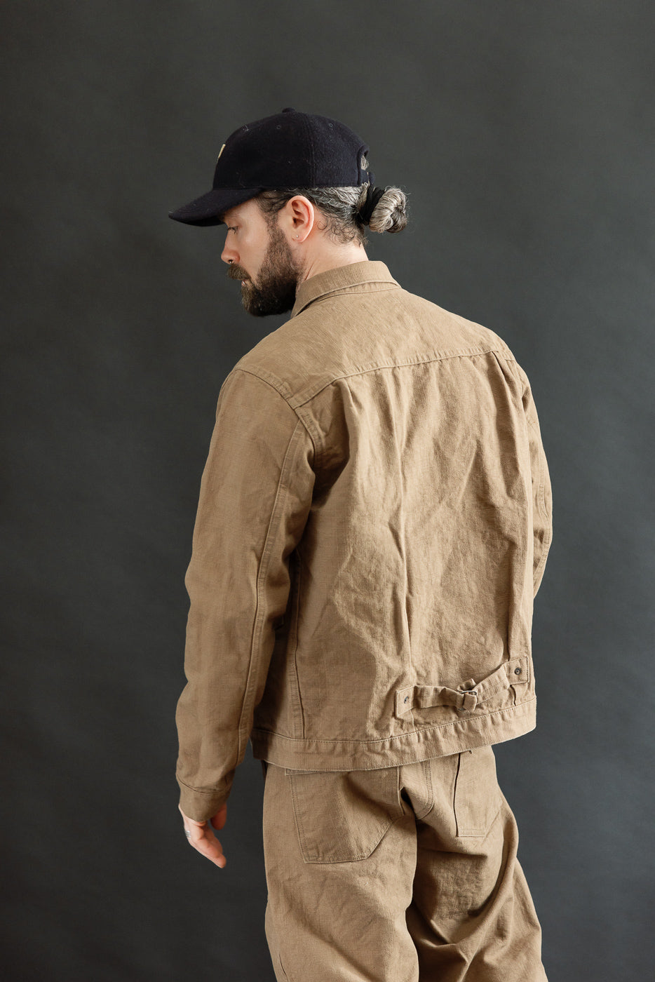 A man models the 01507-HOX-BRK - 13oz Brown Khaki Sulfur Dyed Heavy Oxford Jacket - Type I, showcasing its cinch back and relaxed fit, with a visible belt-style adjuster.