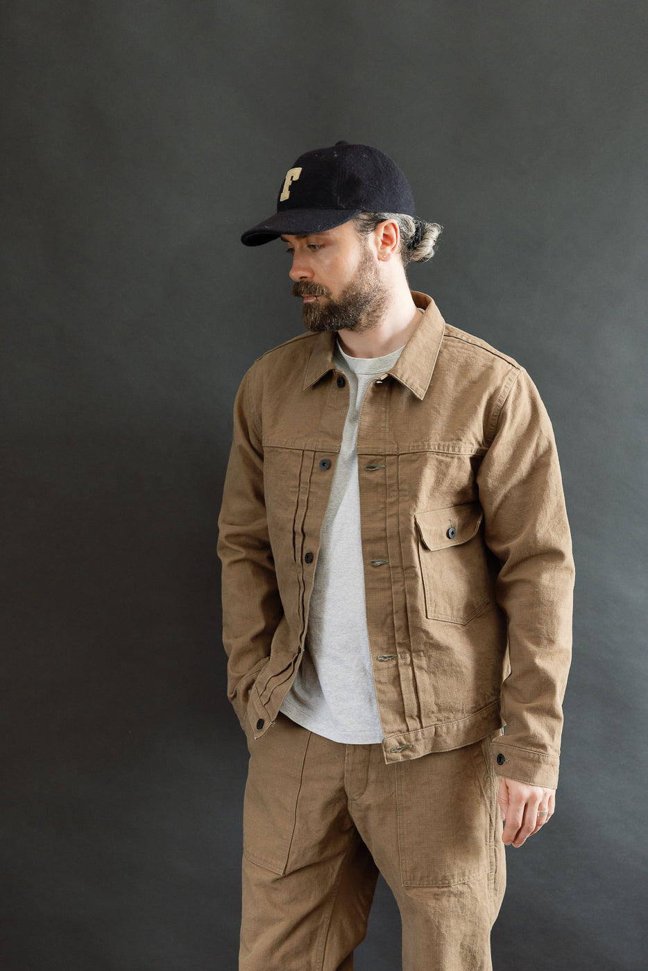 A bearded man models the 01507-HOX-BRK - 13oz Brown Khaki Sulfur Dyed Heavy Oxford Jacket - Type I, showcasing its classic workwear style with buttoned flap pockets and a cinch back.