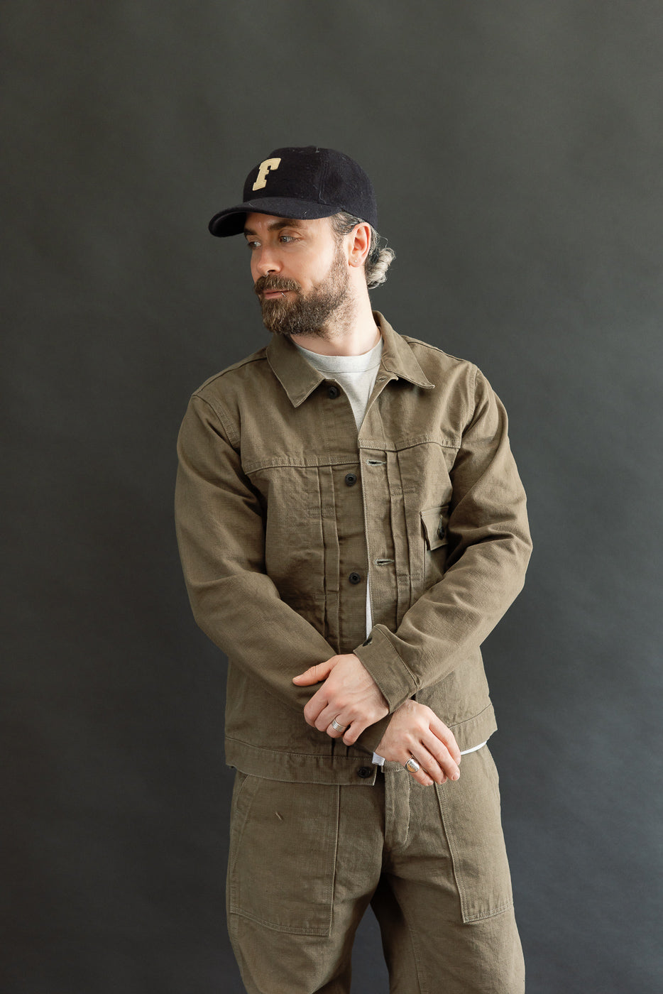 Bearded man models the 01507-HOX-OLB - 13oz Olive Khaki Sulfur Dyed Heavy Oxford Jacket - Type I, showcasing its pleated front, copper rivets, and single chest pocket, reflecting Dant's workwear style.