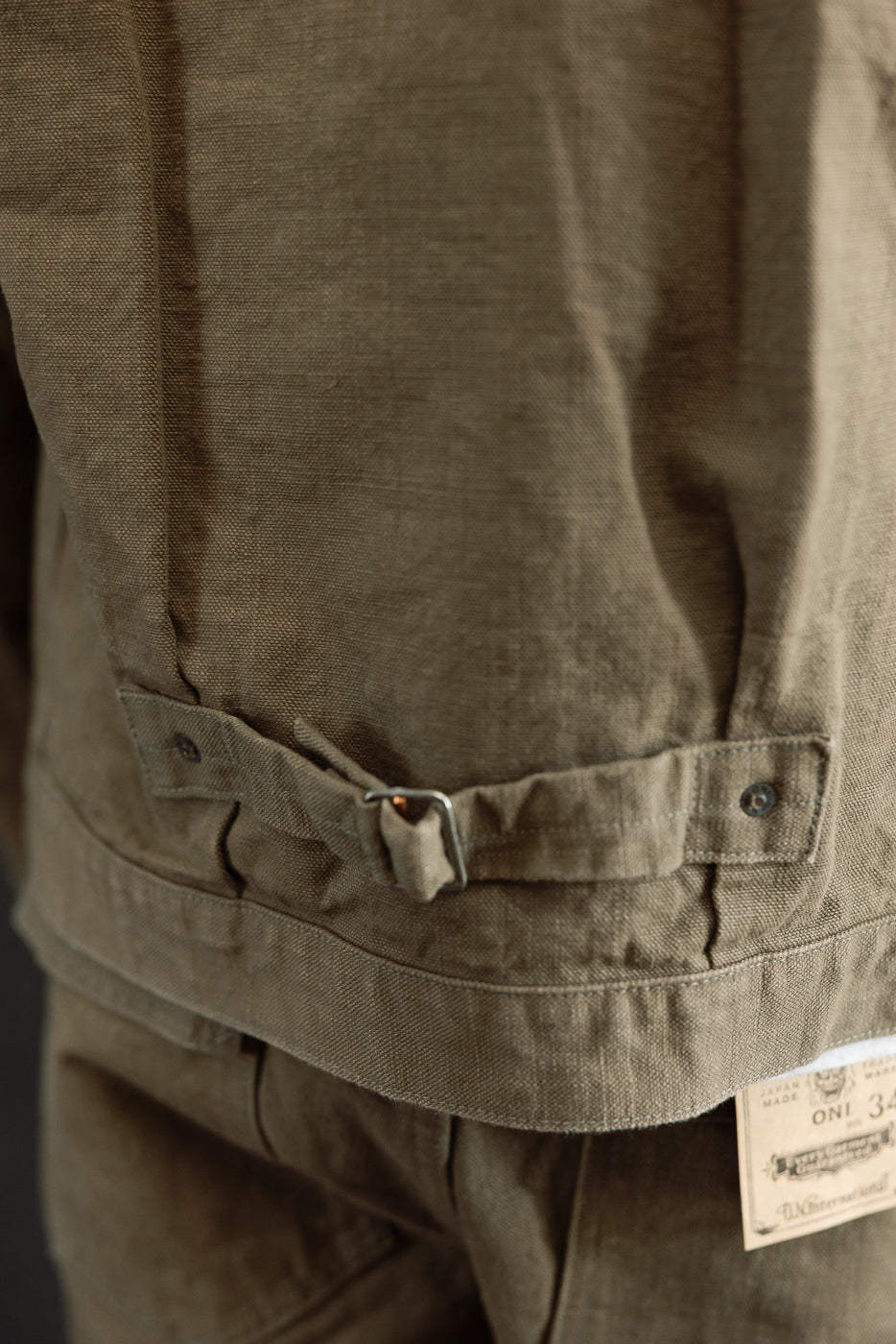 Close-up of the ONI 01507-HOX-OLB jacket's back, highlighting the olive-drab cinch-strap with metal buckle and ONI 34 tag, showcasing its workwear-inspired heritage craftsmanship.