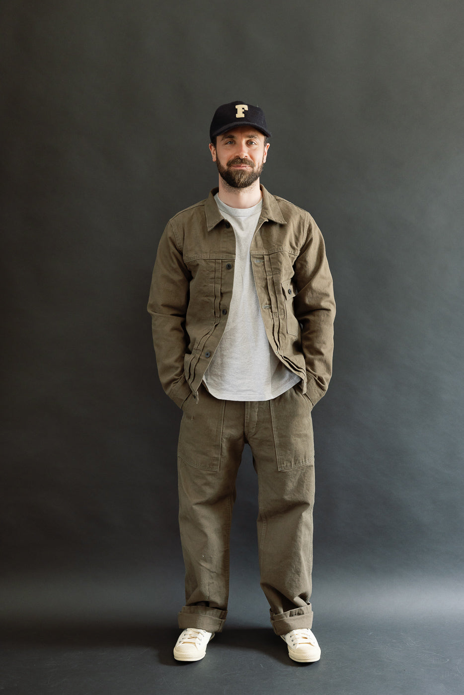 The 01507-HOX-OLB jacket, worn by a casually dressed man, showcases olive khaki sulfur-dyed oxford fabric, featuring classic workwear details like pleats, copper rivets, and a cinch back.