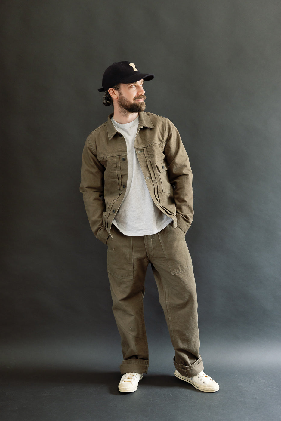 A man models the 01507-HOX-OLB - 13oz Olive Khaki Sulfur Dyed Heavy Oxford Jacket - Type I, showcasing its unique texture and classic workwear silhouette against a plain backdrop.