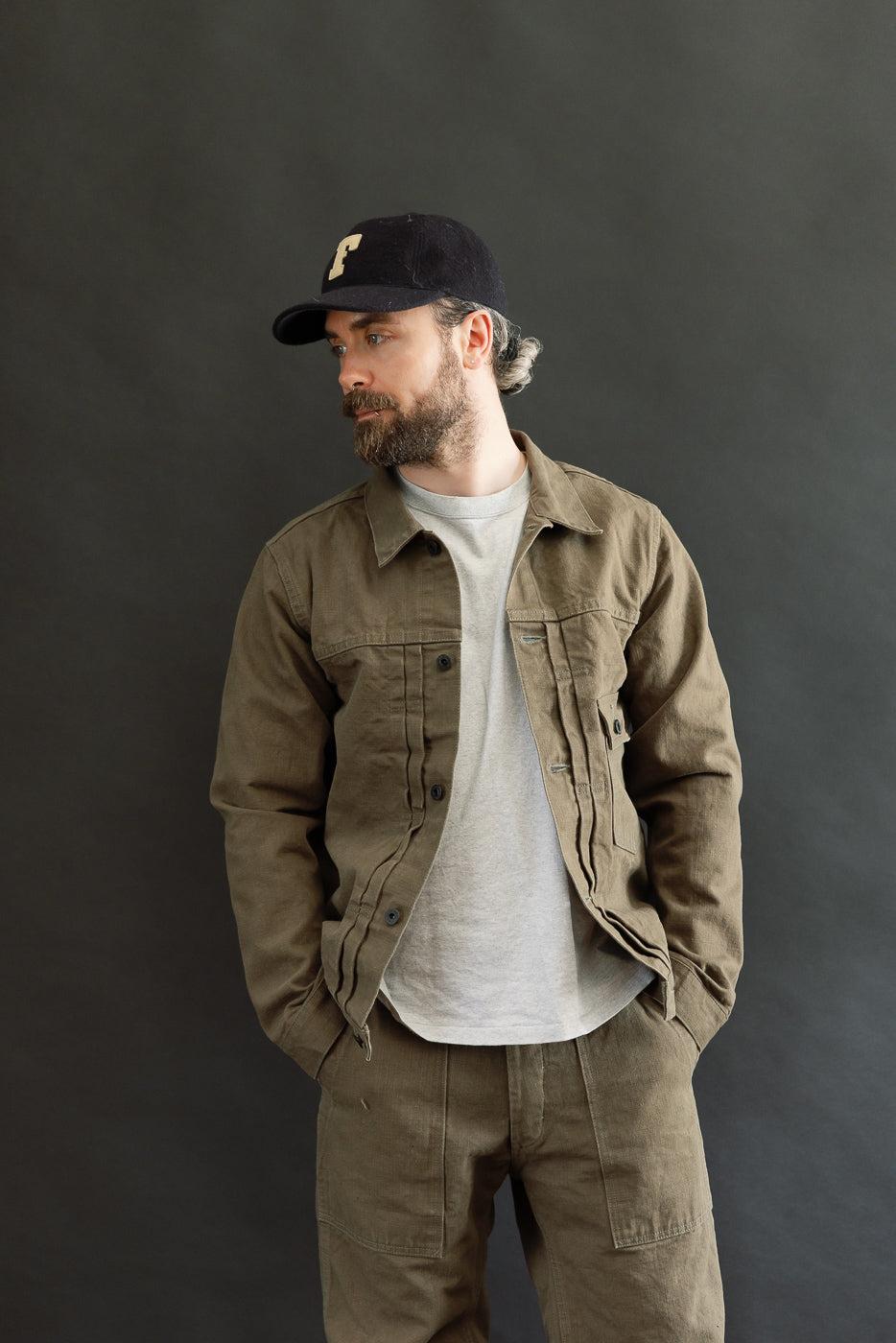 A bearded man models the 01507-HOX-OLB - 13oz Olive Khaki Sulfur Dyed Heavy Oxford Jacket - Type I, showcasing its utilitarian design with pleats, copper rivets, and a single chest pocket.