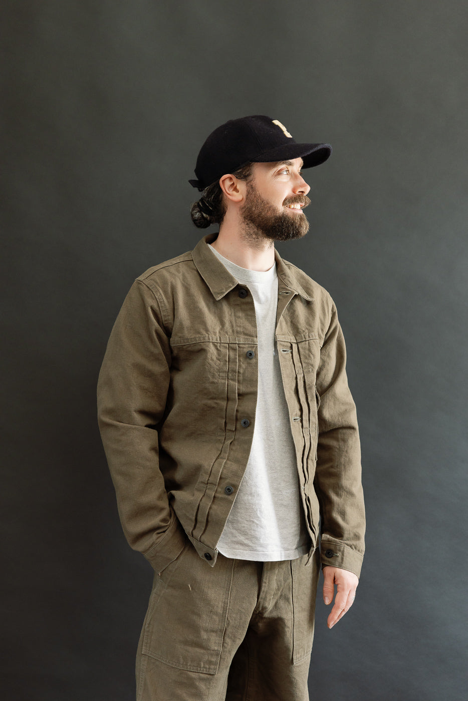 A bearded man models the 01507-HOX-OLB 13oz Olive Khaki Sulfur Dyed Heavy Oxford Jacket - Type I, showcasing its fitted silhouette with double pleats, a left chest pocket, and copper rivets.