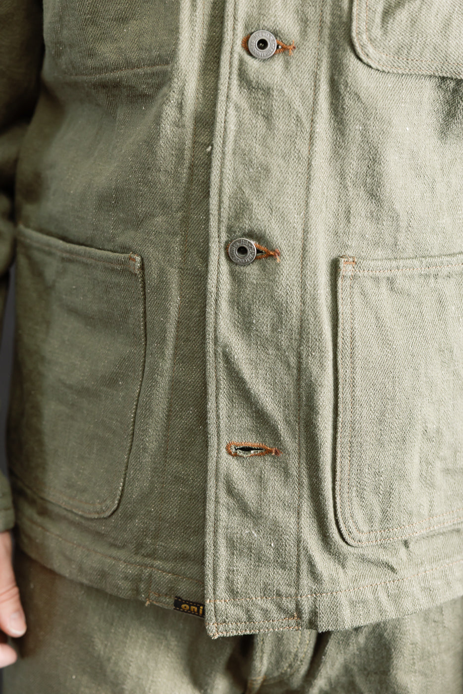 Close-up front view of olive selvedge denim jacket on male model, showing pockets, buttons, and fabric texture.