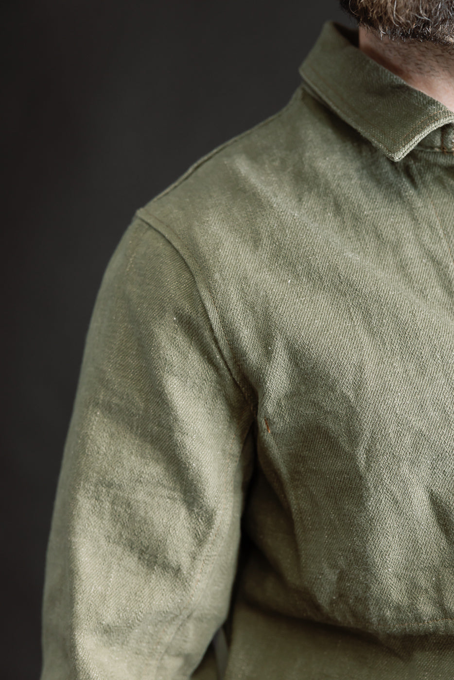 Close-up of male model's upper front left shoulder and collar on olive green selvedge denim jacket, showing fabric texture. Model is 5'9