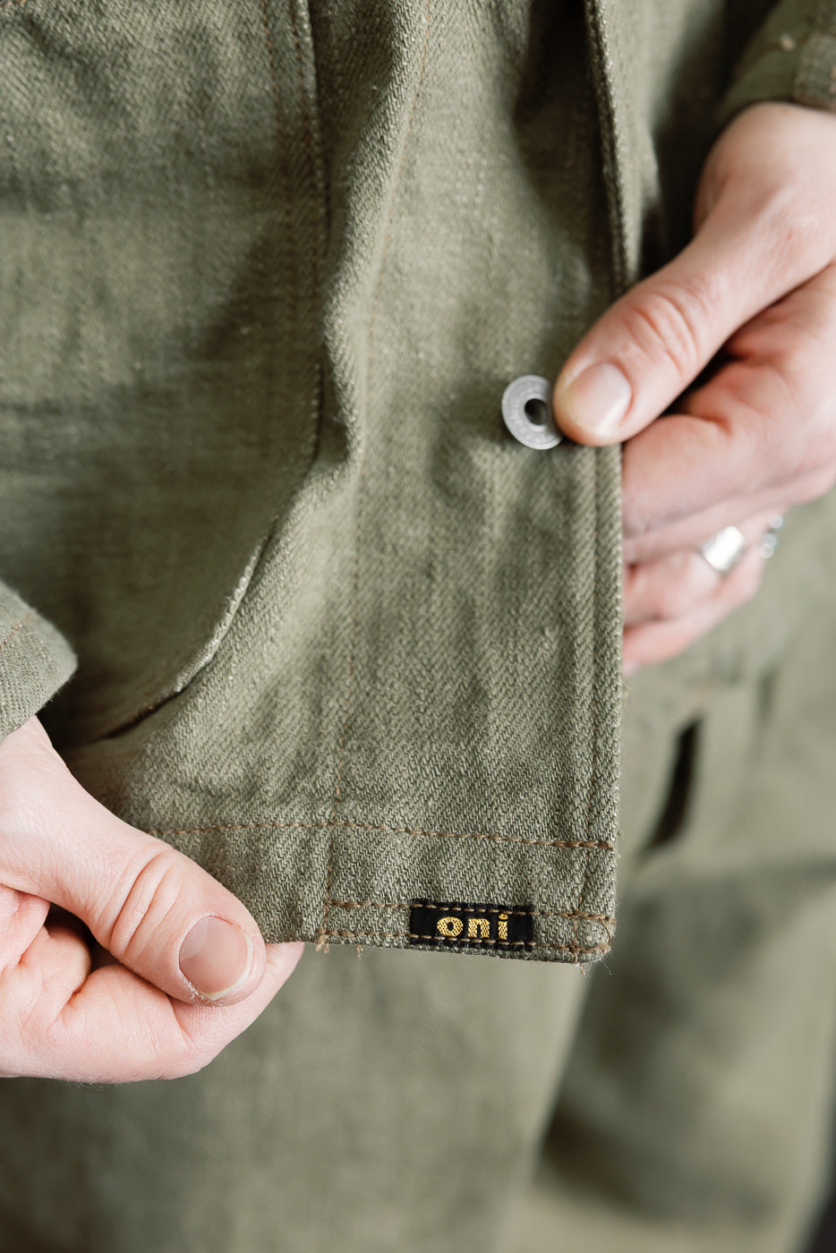 Close-up of front hem and button of olive green selvedge denim jacket, showing fabric texture and label, modeled by a male, 5'9