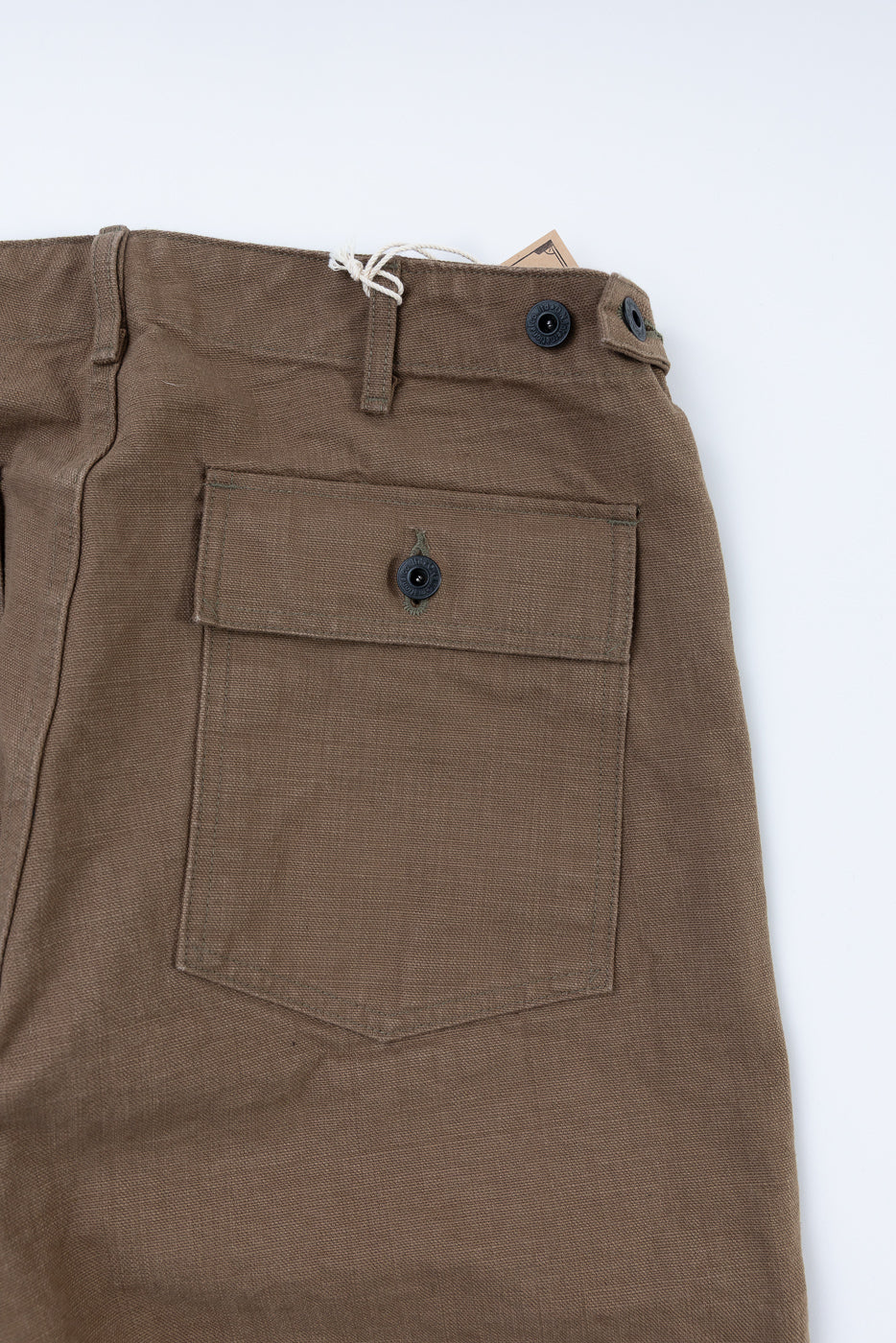 Close-up of 207-HOX-BRK - 13oz Brown Khaki Sulfur Dyed Heavy Oxford Baker Pants, showcasing the waistband with belt loops, button closure, and a button-fastened patch pocket, highlighting its textured, uneven fabric.