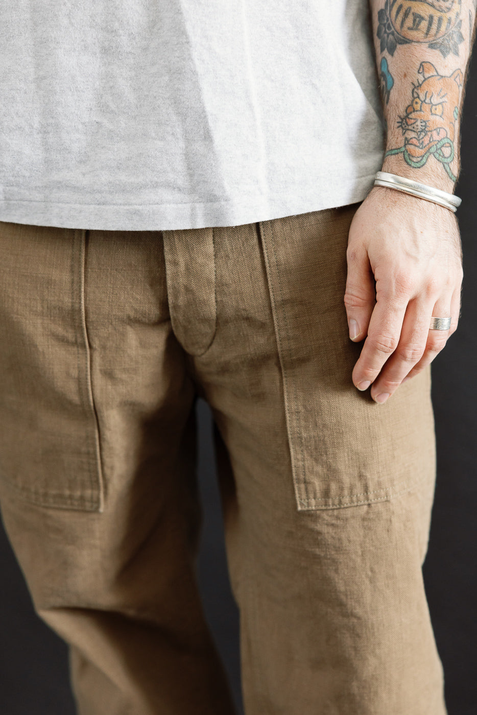 Close-up of the 207-HOX-BRK Brown Khaki Sulfur Dyed Heavy Oxford Baker Pants, showcasing their straight-leg cut and patch pockets, paired with a light gray T-shirt and silver accessories.