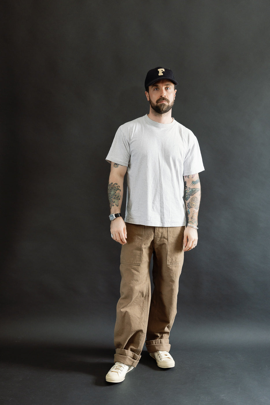 Adult male model wearing 207-HOX-BRK - 13oz Brown Khaki Sulfur Dyed Heavy Oxford Baker Pants, showcasing wide straight cut. Styled with gray T-shirt and off-white sneakers, reflecting Dant's premium menswear aesthetic.