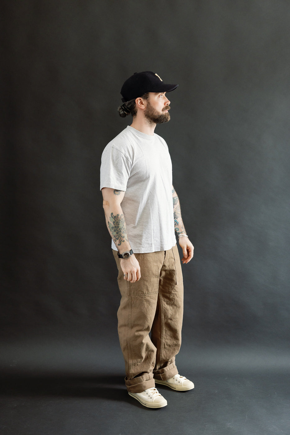 A man models the 207-HOX-BRK - 13oz Brown Khaki Sulfur Dyed Heavy Oxford Baker Pants, showcasing their wide straight cut and relaxed fit, with distinctive textured fabric and cuffed ankles.