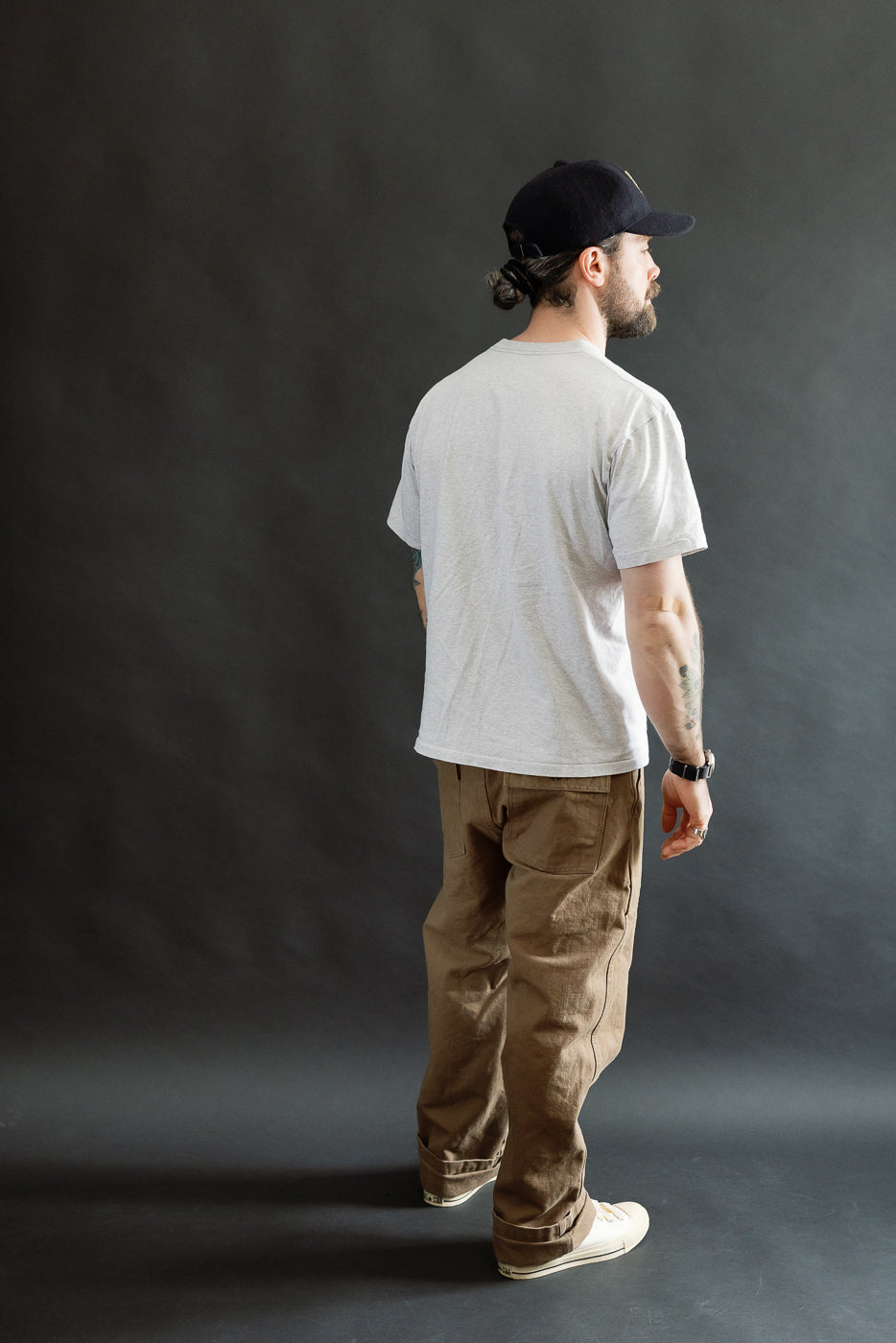 Man in profile wearing ONI Denim 207-HOX-BRK - 13oz Brown Khaki Sulfur Dyed Heavy Oxford Baker Pants, showcasing wide straight cut and relaxed style, paired with light gray T-shirt and canvas sneakers.