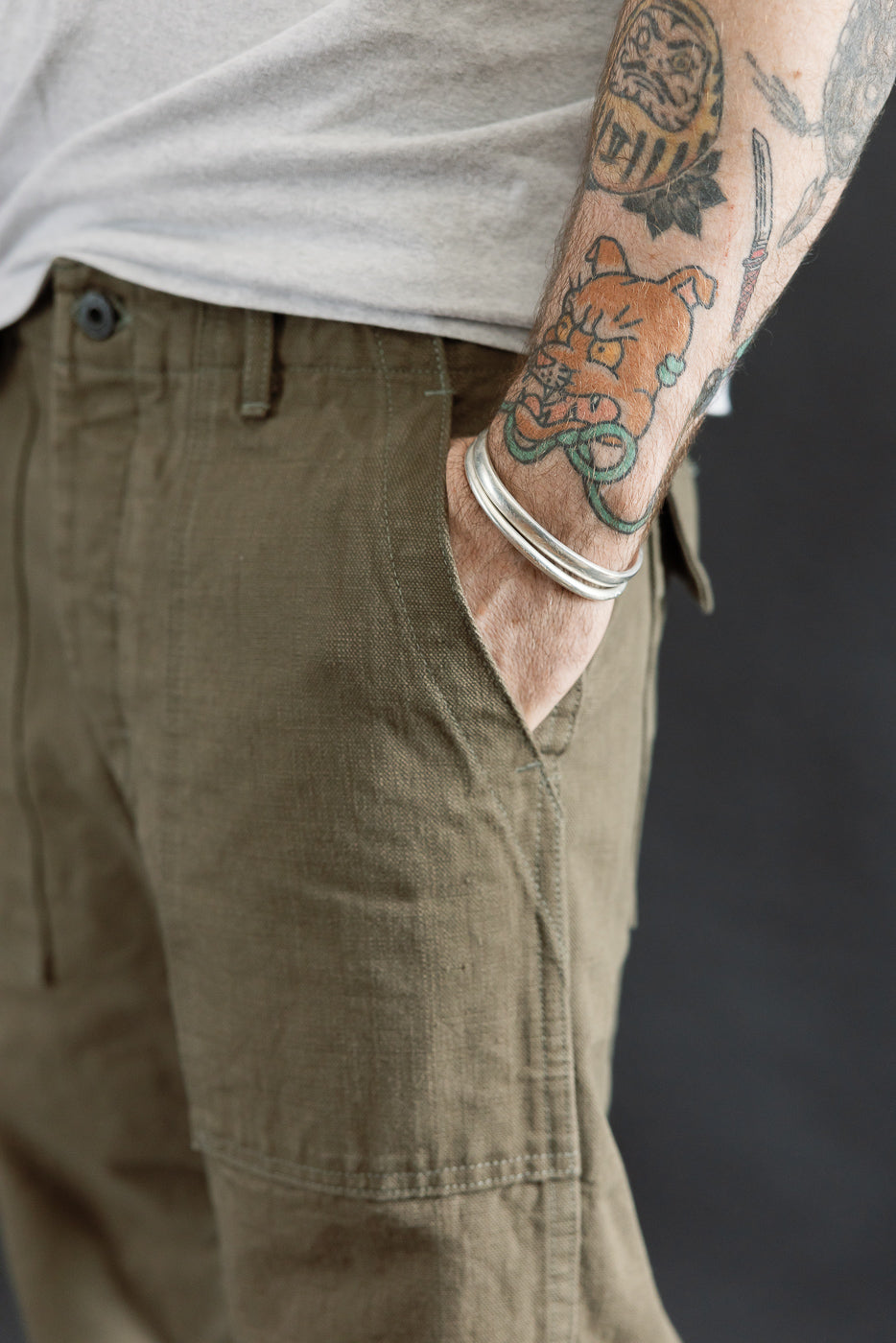 Close-up of the 207-HOX-OLB - 13oz Olive Khaki Sulfur Dyed Heavy Oxford Baker Pants, showcasing their unique texture and relaxed fit, with a hand tucked casually into the pocket.