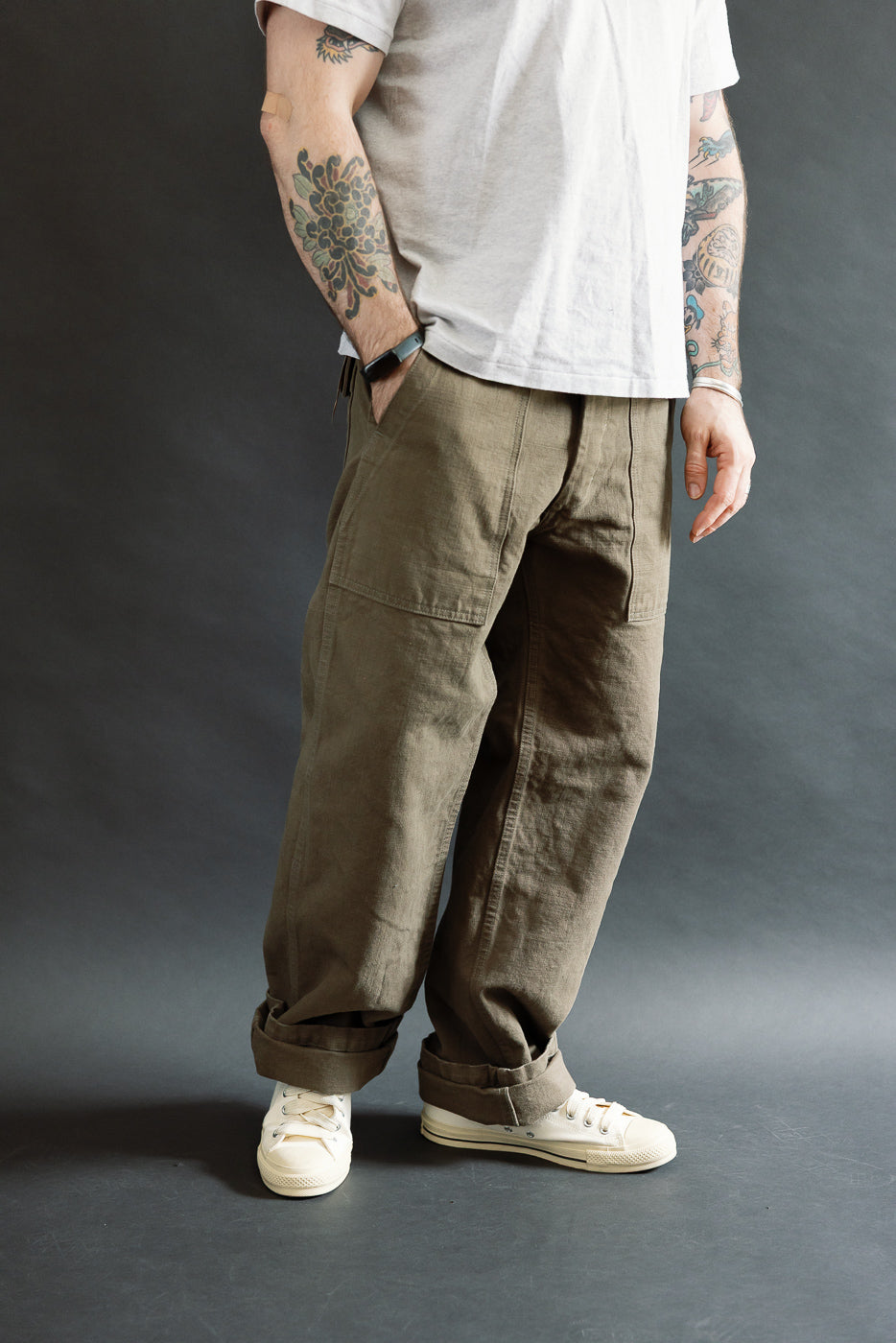 A person showcases the ONI Denim 207-HOX-OLB Olive Khaki Sulfur Dyed Heavy Oxford Baker Pants, featuring a wide straight cut and large patch pockets, styled with off-white canvas sneakers.