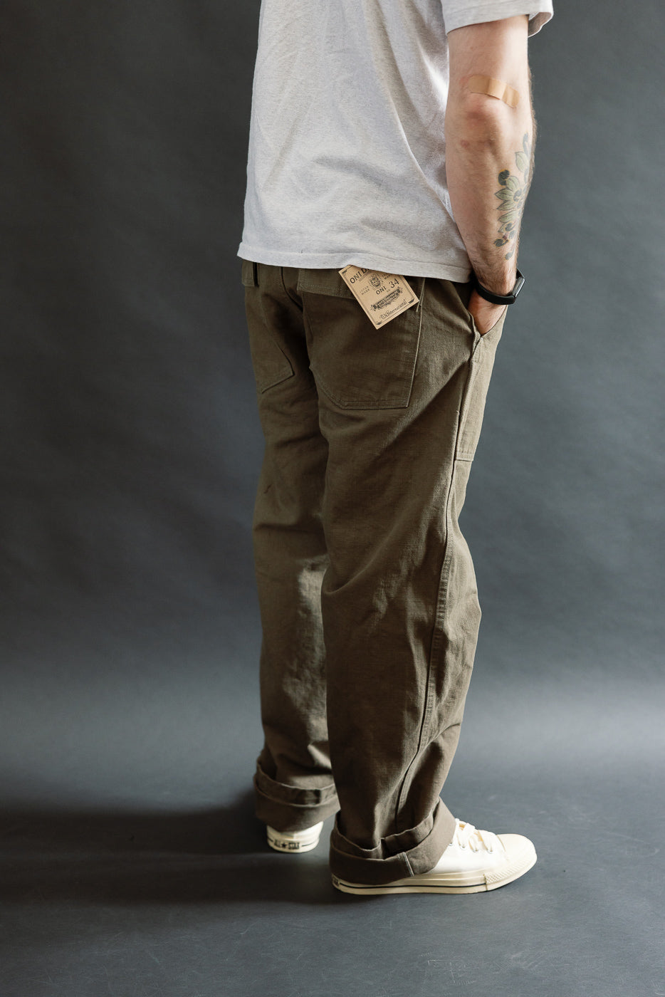 The 207-HOX-OLB Olive Khaki Baker Pants feature a high-rise, wide straight cut, visible paper tag, and unique texture, paired with white canvas sneakers against a minimalist backdrop.