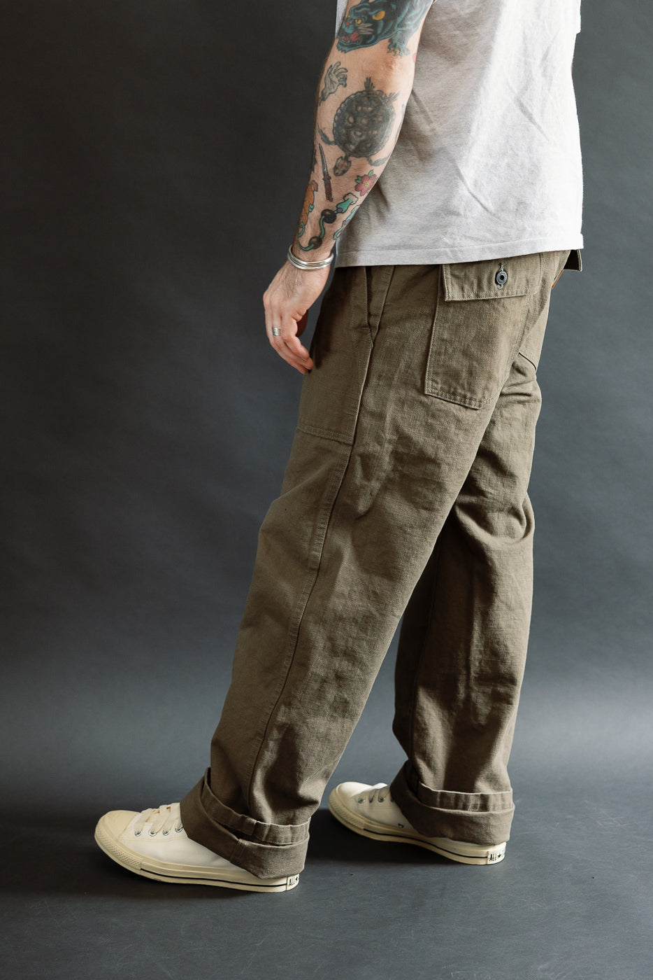 Person wearing 207-HOX-OLB Olive Khaki Sulfur Dyed Heavy Oxford Baker Pants with patch pockets and rolled cuffs, paired with off-white canvas sneakers. The pants feature a relaxed, wide straight cut.
