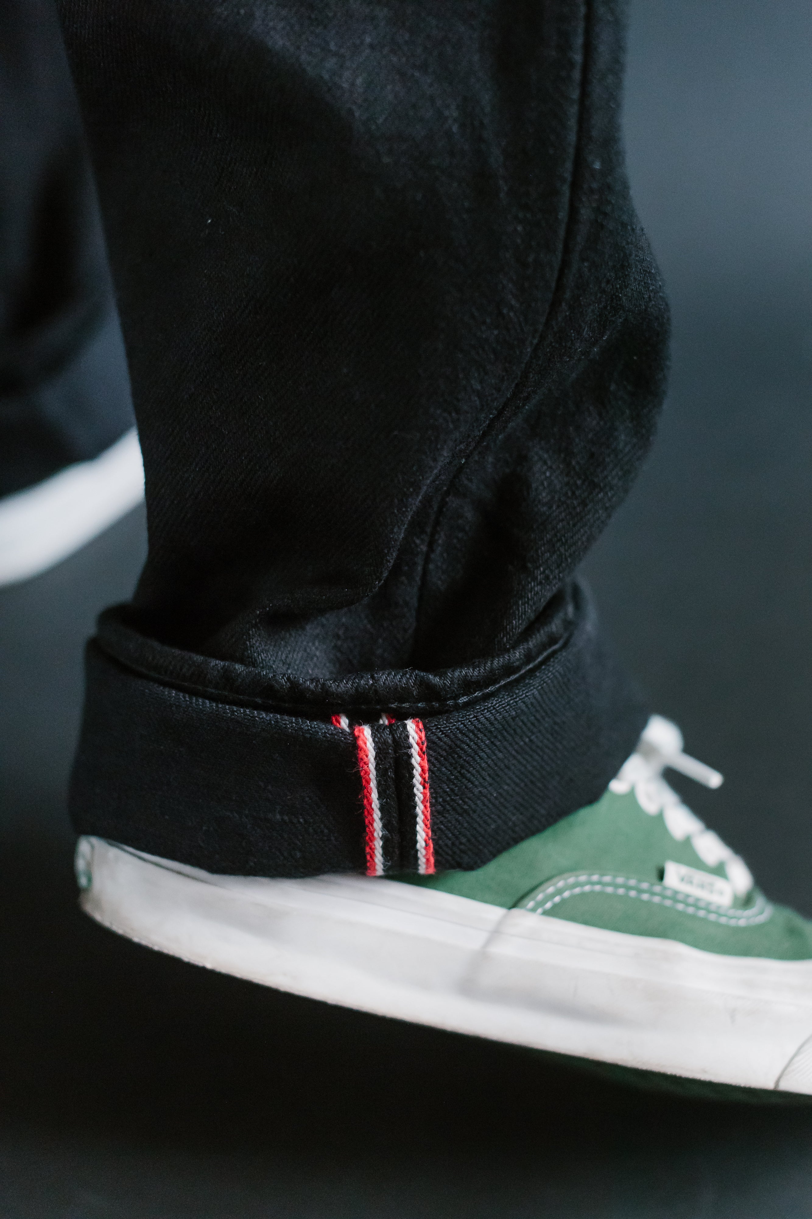 Close-up of black selvedge jeans hem with red/white stripe detail, worn by male model, paired with green sneakers