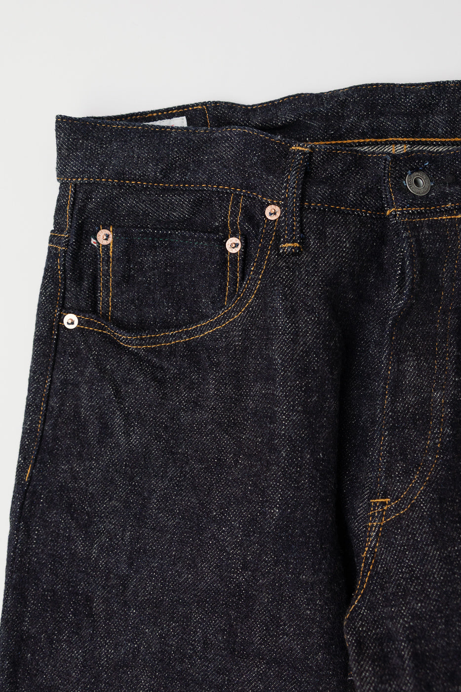Close-up of front left side of dark indigo selvedge denim jeans, showing pocket detail and copper rivets.