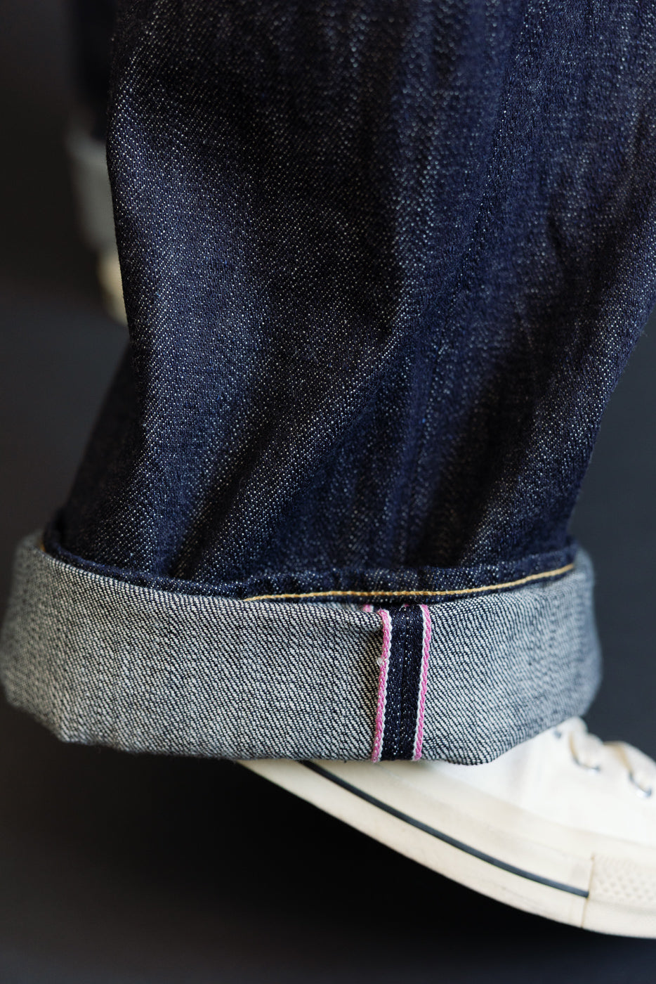 Close-up of male model's rolled denim hem showing selvedge edge, pink stitching, and raw fabric texture with white sneaker
