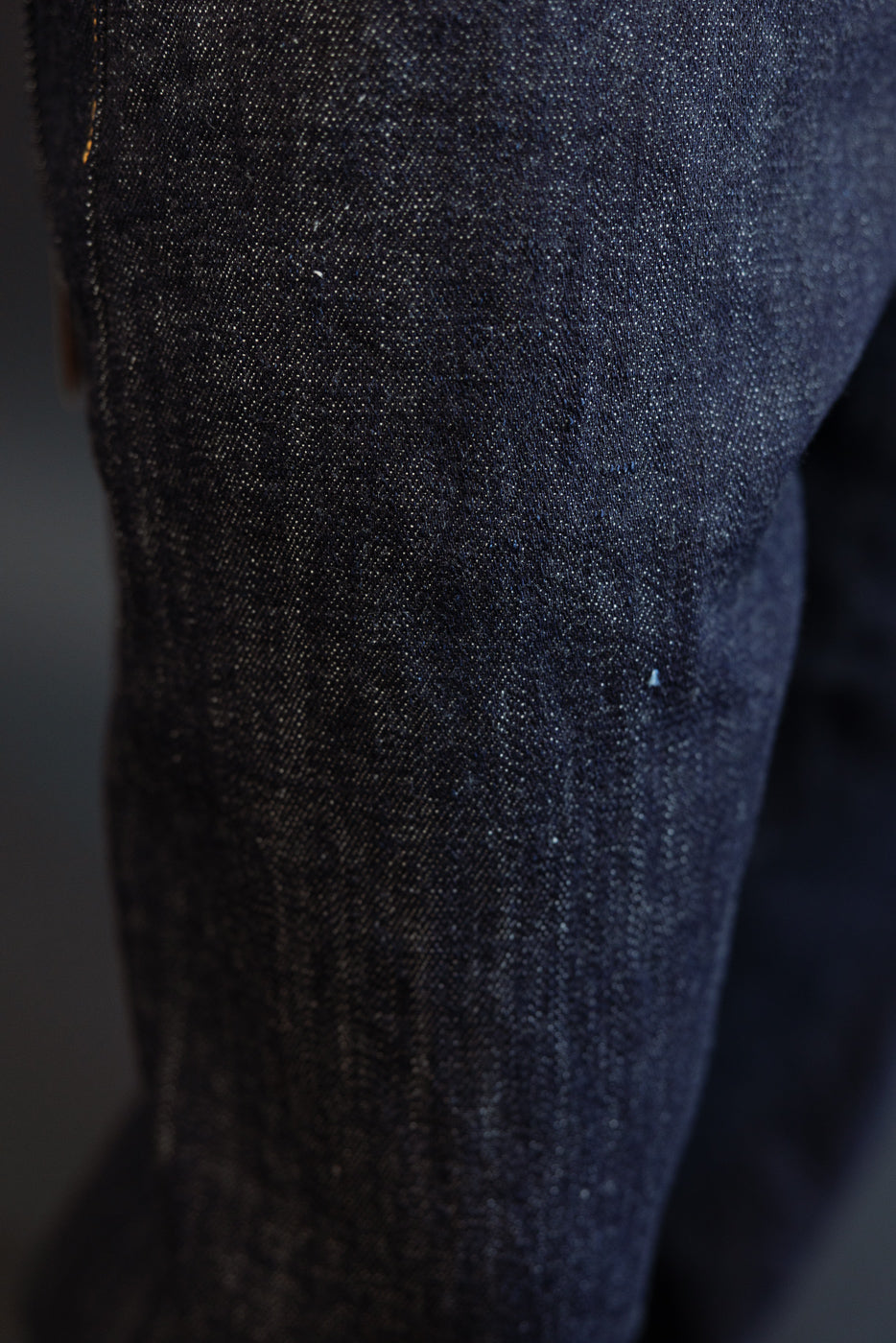 Close-up of dark selvedge denim jeans leg, showcasing fabric texture and weave detail on male model.