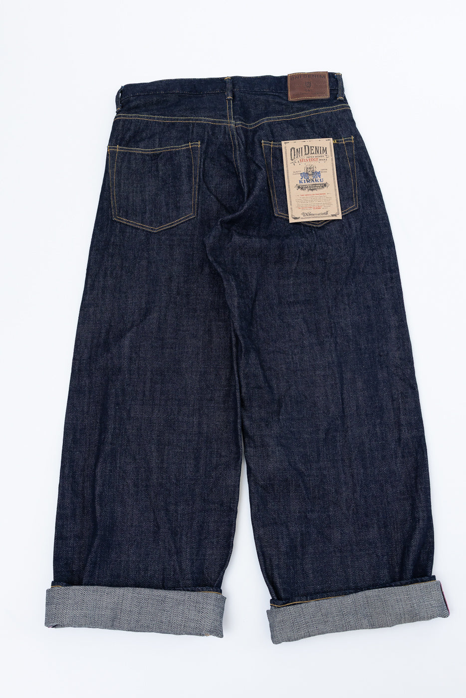 Back view flat lay of dark selvedge denim pants with cuffed hems, branded patch on right pocket, and leather waist tag