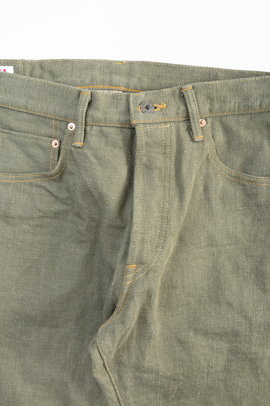 Close-up of front upper section of olive selvedge denim jeans, button fly, coin pocket, and gold stitching.