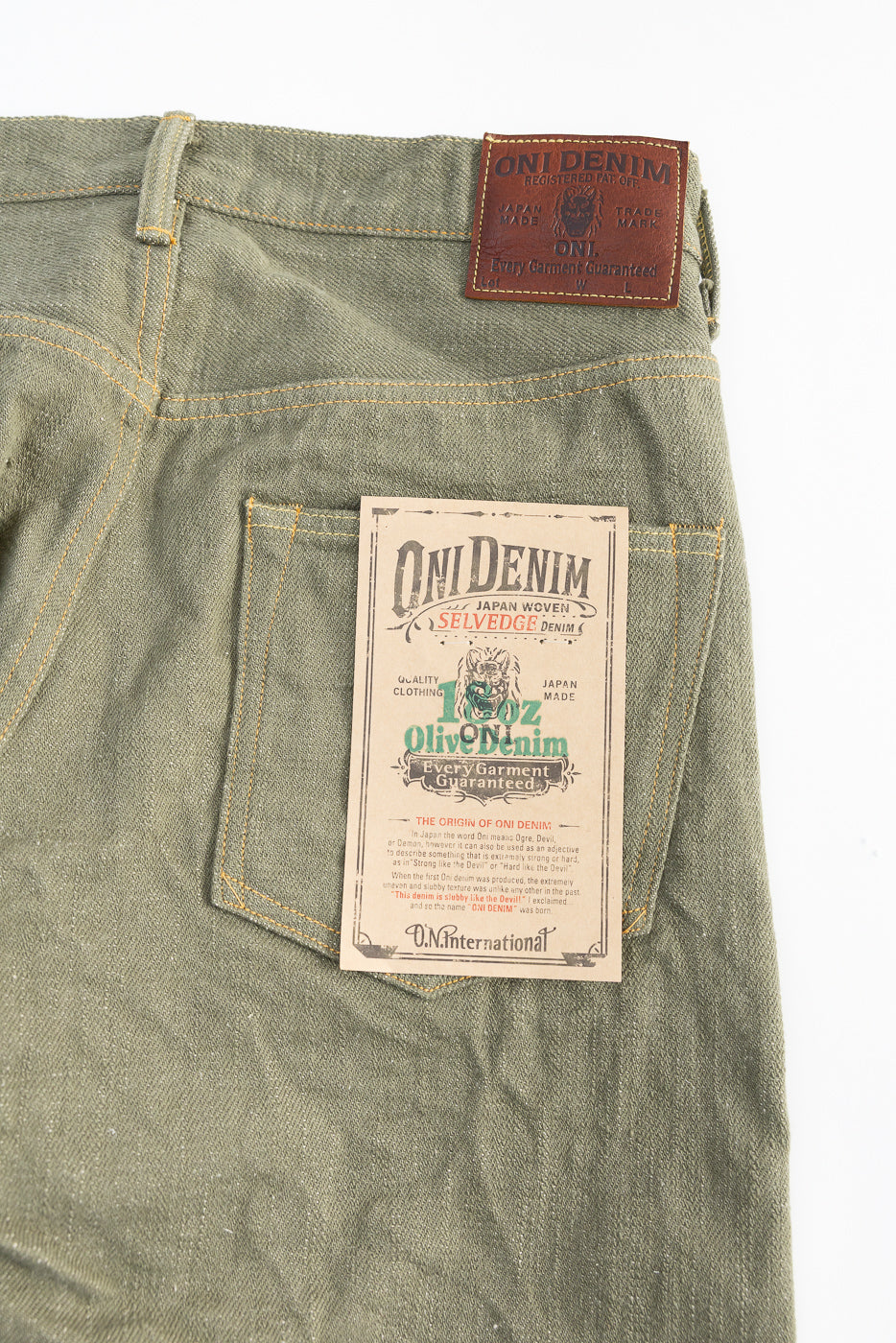 Close-up of back side of olive selvedge denim jeans showing rear pocket, leather patch, and tag details.