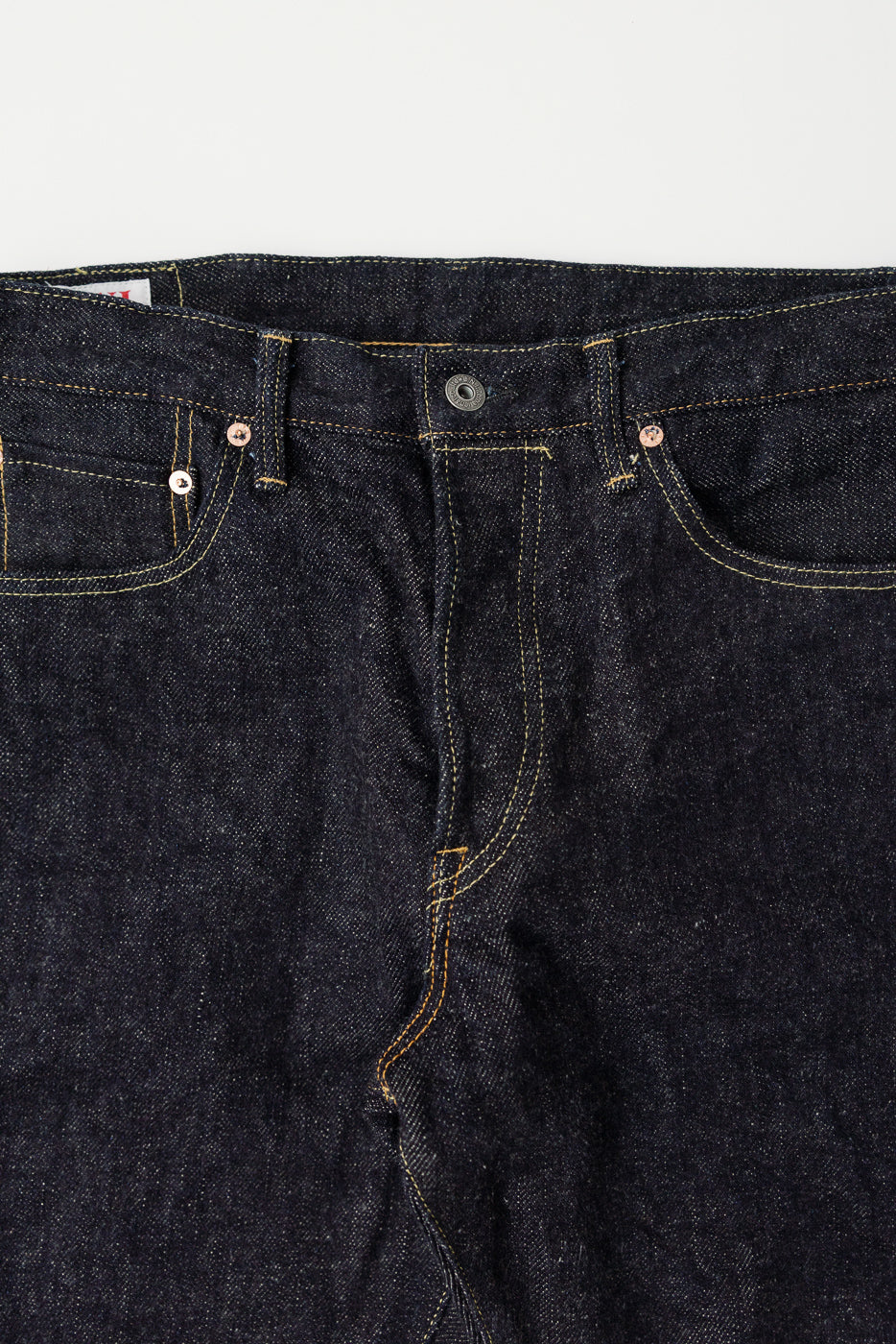 Close-up front view of dark indigo selvedge denim jeans, highlighting top button, rivets, and pocket stitching at Dant.