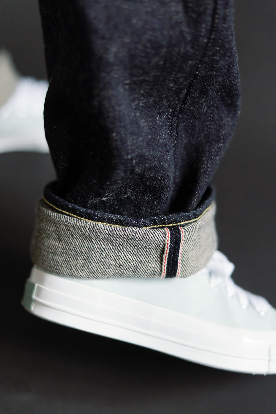 Close-up of ONI 20oz dark indigo selvedge jeans cuff, showing selvedge ID and denim texture on male model's leg, side view above white sneaker.
