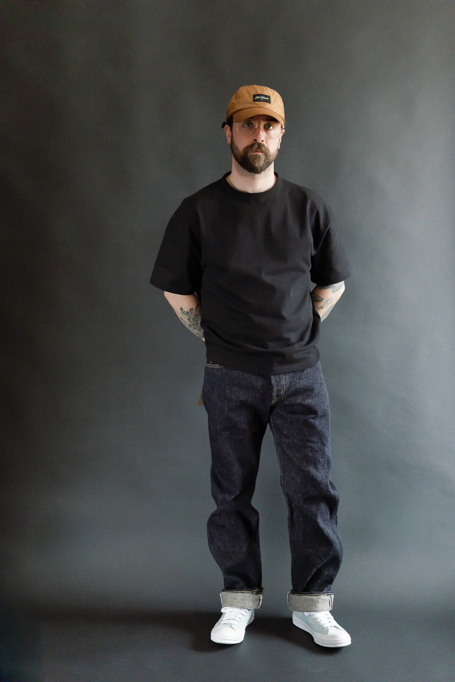 Full body front view of male model 5'9, 165lbs, athletic build wearing Oni 288 DIZR 20oz dark indigo secret denim selvedge jeans, straight fit, cuffed hem, styled with casual black tee and tan cap, Dant contemporary casual apparel.