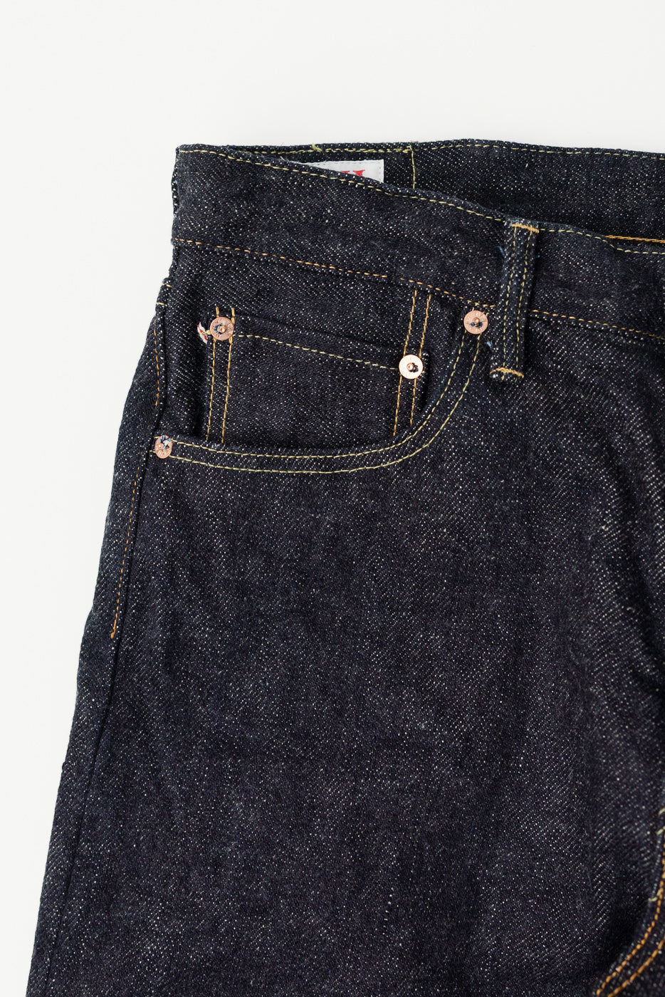 Close-up of front left pocket and waistband on dark indigo selvedge denim jeans, showing copper rivets and textured fabric.