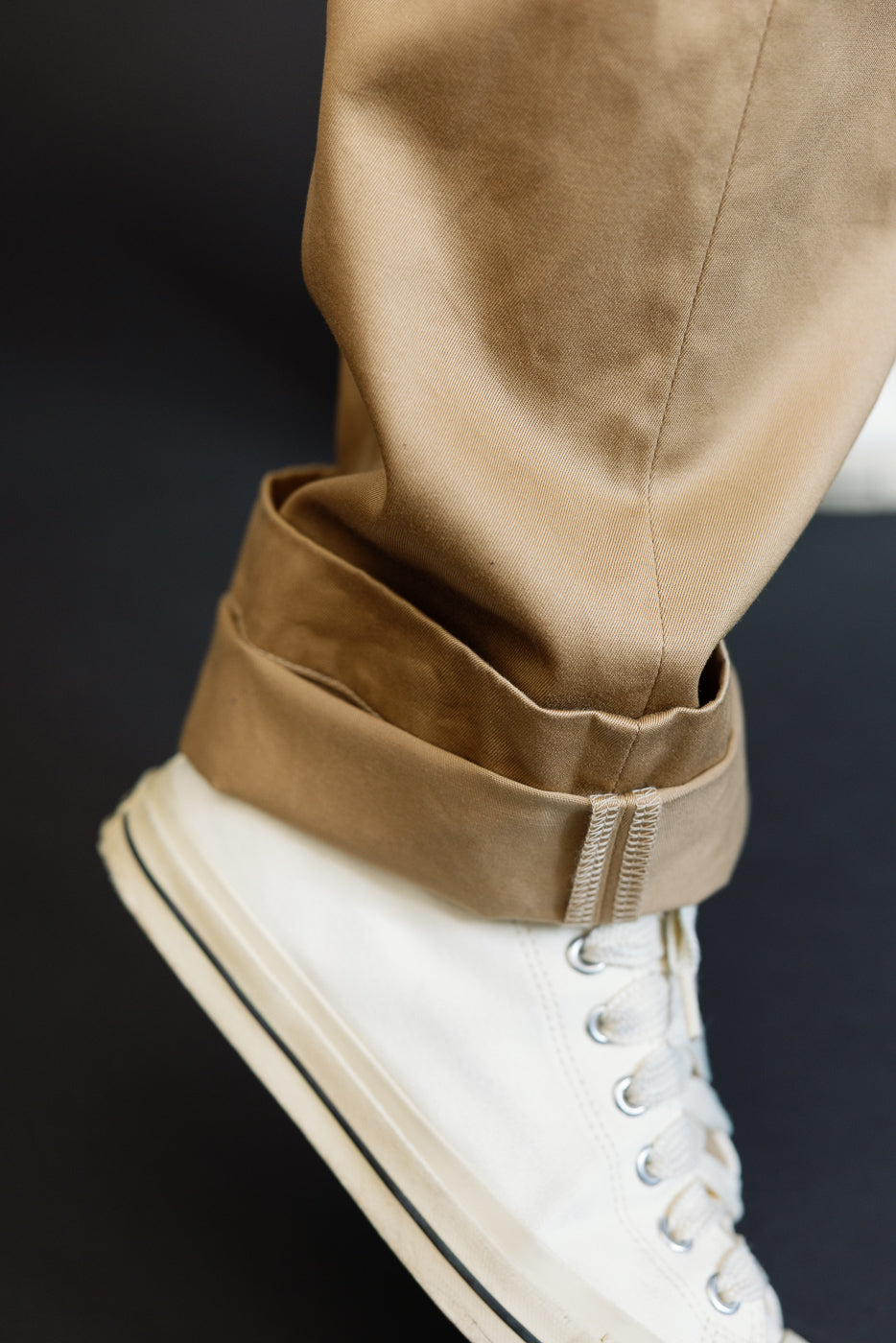 Close-up side view of male model's cuffed khaki pants hem with visible stitching, paired with white high-top sneakers.