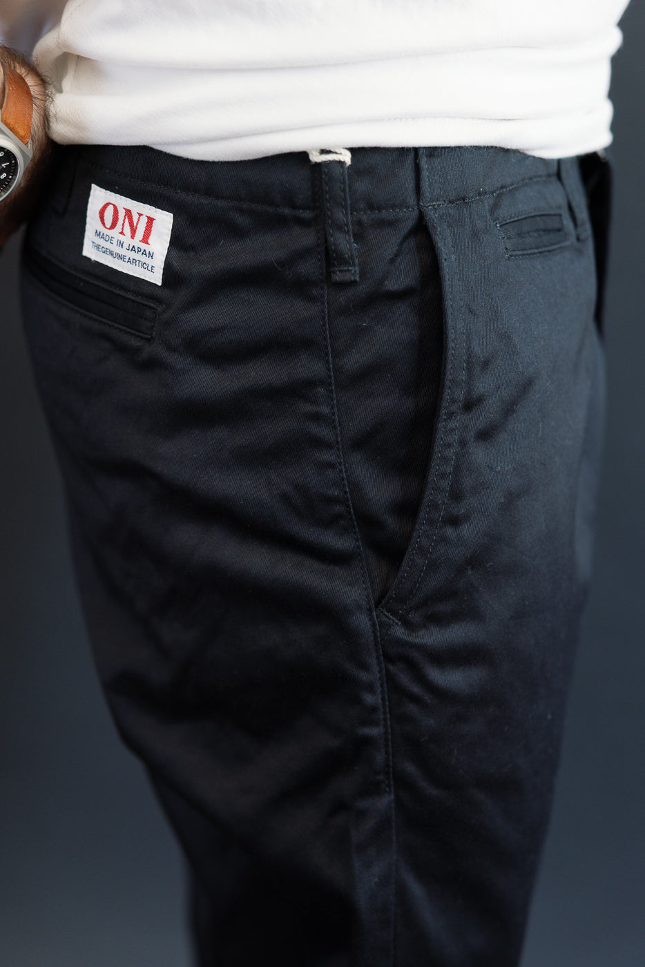Close-up back view of men's dark chino pants, showing ONI brand patch, waistband, and rear pocket detail.