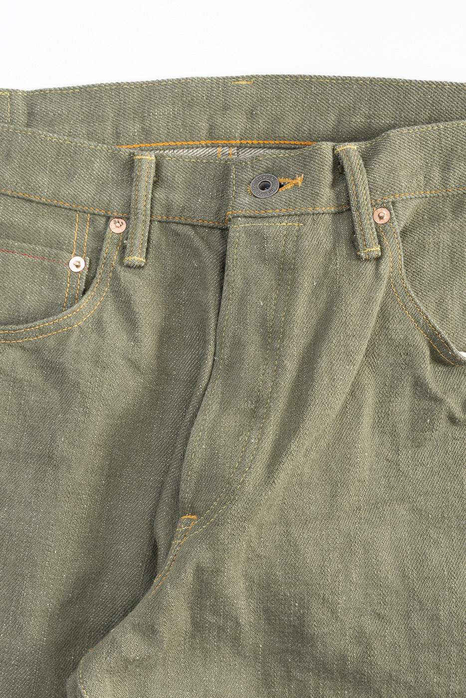 Close-up of front waist and pocket details on olive green selvedge denim jeans, showing button, rivets, belt loops, and textured fabric.